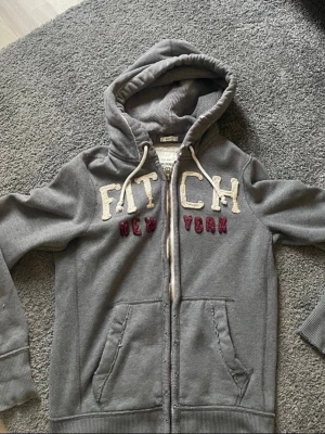 Grå hoodie med dragkedja och brodyr - Säljer en grå hoodie från Abercrombie & Fitch med dragkedja, huva och vita dragsnören. Broderad text 'FITCH' i vitt och 'NEW YORK' i vinrött framtill. Två fickor och ribbade muddar. Perfekt för chill dagar och streetwear vibes.