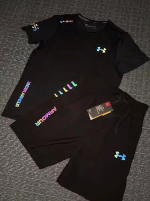 Svart träningsset från Under Armour - Säljer en svart träningsset från Under Armour med färgglada loggor och textdetaljer. Shortsen har snörning i midjan och är tillverkade i ett lätt och sportigt material som passar perfekt för träning. Snygg och modern design med regnbågsfärgade detaljer. Storlek M, helt ny 