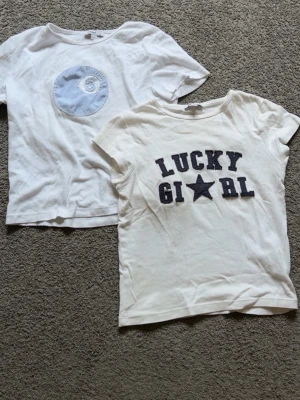 2 vita t-shirts med tryck, storlek S - Två vita t-shirts i bomull, båda med korta ärmar. Ena har ett blått cirkeltryck framtill och den andra har texten 'LUCKY GIRL' i svart med en stjärna. Perfekta till jeans eller shorts för en avslappnad look.