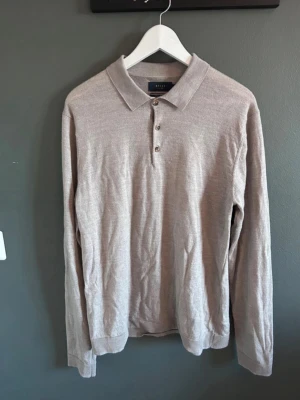 Beige merinoullströja från Riley - Snygg beige långärmad tröja från Riley i fin merinoull. Klassisk pikékrage med tre knappar framtill och ribbade muddar vid ärmslut och nederkant. Perfekt för dig som gillar stilrena och bekväma plagg.