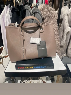Taupe Emporio Armani shoppingväska - Snygg shoppingväska från Emporio Armani i taupefärgat, strukturerat läder. Väskan har både handtag och en bred axelrem med logotyp, samt silverfärgade metalldetaljer. Perfekt storlek för att få plats med allt du behöver. Stängs med dragkedja och har en stilren look.