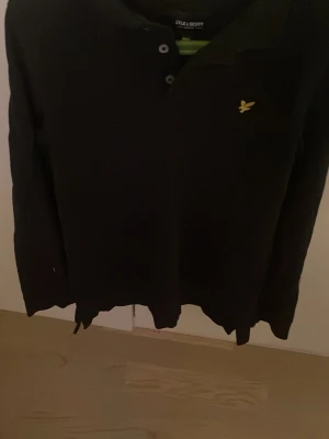 Svart långärmad tröja Lyle & Scott - Klassisk svart långärmad tröja från Lyle & Scott med knappslå och den ikoniska gula loggan broderad på bröstet. Tröjan har en rak passform och är tillverkad i ett mjukt material som passar perfekt till en avslappnad stil.