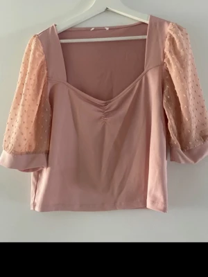 Ljusrosa topp med puffärmar - Superfin ljusrosa topp med transparenta puffärmar i mesh och diskreta broderade detaljer. Toppen har en lätt rynkning framtill och fyrkantig urringning. Perfekt för dig som vill ha en söt och trendig look.