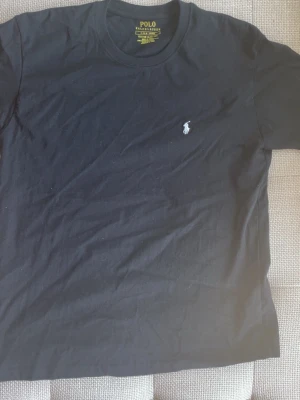 Svart t-shirt från Polo Ralph Lauren - Klassisk svart t-shirt från Polo Ralph Lauren med den ikoniska broderade loggan på bröstet. T-shirten har rund hals och korta ärmar, perfekt till jeans eller shorts. Tillverkad i mjuk bomull för skön känsla.