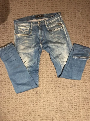 Replay Anbass Jeans - Snygga blå slim fit jeans från Replay med slitna detaljer och ljusare partier framtill och baktill. Klassisk femficksmodell med vita sömmar och Replay-logga på bakfickan. Perfekta för en avslappnad och trendig look. Storlek W33 L34!