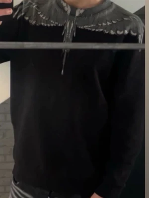 Svart hoodie med grått fågeltryck - Säljer en svart hoodie med ett unikt grått fågelmönster runt halsen och ner över bröstet. Hoodien har lång ärm och en relaxed passform, perfekt för dig som vill sticka ut med en cool detalj. Passar till det mesta och är riktigt bekväm.