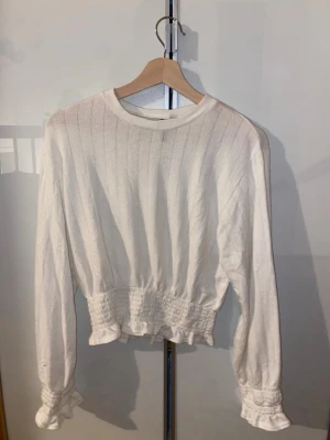 Vit croppad tröja med volangkant - Säljer en vit långärmad croppad tröja med smockad midja och volangkant nertill samt vid ärmsluten. Tröjan har diskreta vertikala ränder och rund halsringning. Perfekt för dig som gillar en söt och trendig stil.