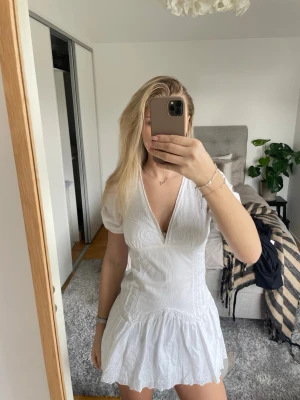 Vit kortklänning med v-ringning - Säljer en vit kortklänning med v-ringning och puffade kortärmar. Klänningen har broderade detaljer upptill och en volang nedtill som ger en feminin look. Perfekt för varma dagar och har en figurnära passning. Materialet känns som bomull och är lätt och luftigt.