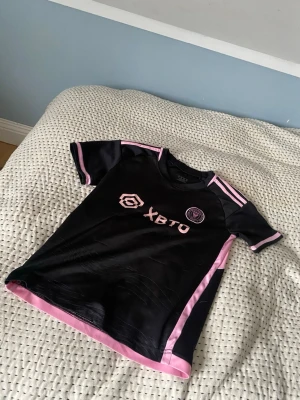 Inter Miami Messi matchtröja svart/rosa - Snygg svart Inter Miami matchtröja från Adidas med rosa detaljer och Messi 10-tryck på ryggen. Tröjan har korta ärmar, klubbmärke på bröstet och sponsorlogga framtill. Tillverkad i lätt och ventilerande material, perfekt för fotbollsträning eller som supporterplagg. Passar till 10-12 år. 