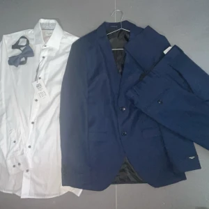 Mörkblå kostym med vit skjorta & fluga - Snygg mörkblå kostym från Selected Homme med matchande kavaj och byxor. Till setet hör en vit skjorta i bomull och en marinblå fluga. Perfekt för dig som vill ha en klassisk och stilren look. Kavajen har två knappar och skjortan har mörka knappar.