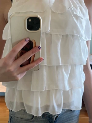 Vit volangtop från H&M - Söt vit ärmlös topp från H&M med flera lager volanger och rund halsringning. Toppen är tillverkad i ett lätt och luftigt material som ger en flowy känsla, perfekt för varma dagar. Stängs med en knapp i nacken.