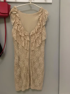 Beige spetsklänning med volang - Supersöt beige kortklänning i spets med volangdetaljer runt axlarna. Klänningen är ärmlös och har ett romantiskt blommigt spetsmönster. Perfekt för dig som gillar en feminin och chic stil.