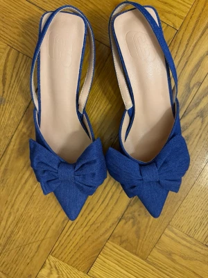 Blå pumps med rosett och låg klack - Snygga blå pumps med spetsig tå och stor dekorativ rosett framtill. Skorna har öppen häl med smal rem och låg klack. Tillverkade i tygmaterial med beige innersula.