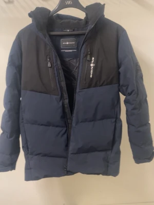 Sail racing jacka  - Snygg blå dunjacka med svarta partier från sail racing. Jackan har två bröstfickor med dragkedja, huva och logga på bröstet samt ryggen. Perfekt för kalla vinterdagar med sin tjocka vaddering och sportiga look.