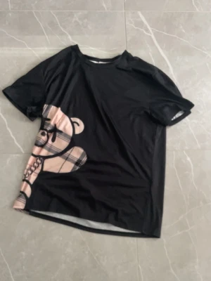 Svart t-shirt med beige mönster - Cool svart t-shirt med korta ärmar och ett stort beige rutigt mönster på framsidan som liknar en björn. T-shirten har en avslappnad passform och är tillverkad i mjukt bomullsmaterial. Perfekt för dig som vill sticka ut med en unik design.