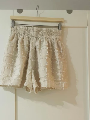 Beige shorts med fransig struktur - Unika beige shorts med hög midja och elastisk midjeresår. Shortsen har ett rutigt, fransigt mönster som ger en cool och trendig look. Perfekta för varma dagar och lätta att matcha med olika toppar.