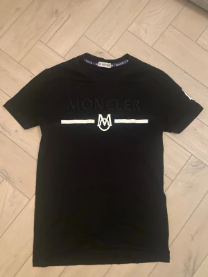 Svart Moncler t-shirt med logga - Snygg svart t-shirt från Moncler med stor logga och vit detalj framtill. Klassisk rund halsringning och korta ärmar. Perfekt för dig som gillar stilrena plagg med exklusiv känsla. Tillverkad i mjuk bomull för skön komfort.