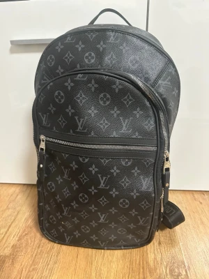 Svart Louis Vuitton ryggsäck - Snygg svart ryggsäck från Louis Vuitton med klassiskt monogrammönster i grått. Väskan har två stora fack med silverfärgade dragkedjor och justerbara axelband. Materialet är slitstark canvas och mesh på baksidan för extra komfort. Perfekt för dig som vill ha en exklusiv look.