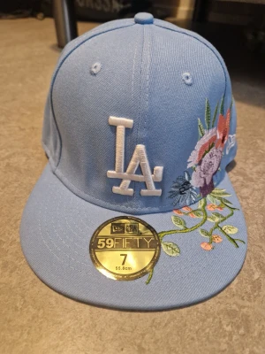 Ljusblå LA-keps med broderade blommor - Snygg ljusblå 59FIFTY-keps från New Era med LA-logga framtill i vitt och broderade blommor i lila, rött och grönt på sidan. Kepsen har rak skärm och klassisk passform. Materialet är bomullstyg som känns både mjukt och slitstarkt.