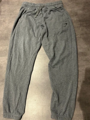 Gråa mjukisbyxor från Gymshark - Säljer ett par gråa mjukisbyxor från Gymshark med dragsko i midjan och resår vid bensluten. Byxorna har en slim fit passform och är tillverkade i ett mjukt och stretchigt material. Perfekta för chill eller träning.