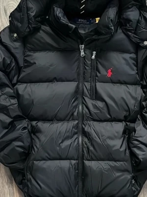 Svart pufferjacka från Polo Ralph Lauren - Snygg svart pufferjacka från Polo Ralph Lauren med huva och dragkedja framtill. Jackan har en röd broderad logga på bröstet och stora quiltade partier. Perfekt för kalla vinterdagar och riktigt trendig look.