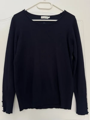 Mörkblå stickad tröja med knappar micha - Mörkblå stickad tröja från MICHA med rund halsringning och diskreta prickdetaljer i stickningen. Tröjan har långa ärmar och dekorativa guldiga knappar längs ryggen och vid ärmsluten. Ribbstickade muddar nedtill och vid ärmarna. Använda fåtal gånger 