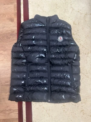Svart glansig dunväst från Moncler - Snygg svart dunväst från Moncler med glansig finish och klassisk Moncler-logga på bröstet. Västen har ståkrage, dragkedja framtill och två sidofickor. Perfekt för lager-på-lager och ger en trendig look till din outfit. Storleken är XL men den passar som M och L