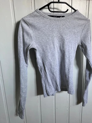 Ljusgrå ribbad långärmad topp - En enkel och stilren ljusgrå långärmad topp från Vero Moda. Toppen är ribbad och har en tight passform med rund halsringning. Perfekt att matcha med jeans eller kjol för en clean look. (Använd max 5 gånger)