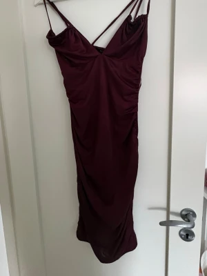 Vinröd klänning med smala band - Snygg vinröd klänning med smala axelband och v-ringad urringning. Klänningen har en figurnära passform och lätt rynkade detaljer längs sidorna. Perfekt för dig som vill ha en elegant och stilren look.