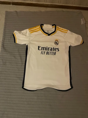 Real Madrid matchtröja Mbappé nr 9 - Vit Real Madrid matchtröja från Adidas med gula och svarta detaljer, tryck av Mbappé och nummer 9 på ryggen. Tröjan har klubbmärke och sponsorlogga framtill samt korta ärmar och sportig passform. Perfekt för fotbollsfans!