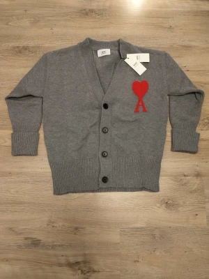 Grå cardigan från Ami Paris - Snygg grå cardigan från Ami Paris med djup V-ringning och tre knappar framtill. Tröjan har ribbade muddar och nederkant samt ett stort rött hjärta med bokstaven A broderat på bröstet. Perfekt för dig som gillar stilrena plagg med en twist.