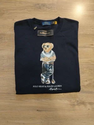 Mörkblå sweatshirt Polo Ralph Lauren - Mörkblå sweatshirt från Polo Ralph Lauren med ikoniska Polo Bear-trycket framtill. Rund halsringning, långa ärmar och ribbade muddar vid ärmslut och nederkant. Tillverkad i mjukt bomullsmaterial för en bekväm känsla.! ”Det står att den är Large men den är i storleken Small”!