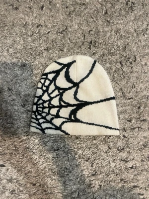 Vit mössa  - Cool vit mössa med svart spindelnätsmönster på sidan. Mössan är stickad och har en mjuk, bekväm passform som passar perfekt till höst och vinter. Perfekt accessoar för dig som vill sticka ut lite extra.