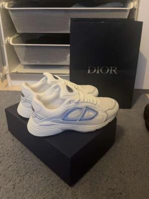 Vita Dior b30 - Snygga vita sneakers från Dior med blå detaljer och tydligt CD-märke på sidan. Skorna har en chunky sula och är tillverkade i mesh och läder för en modern och sportig look. Perfekta för dig som vill sticka ut med exklusiva och trendiga sneakers.