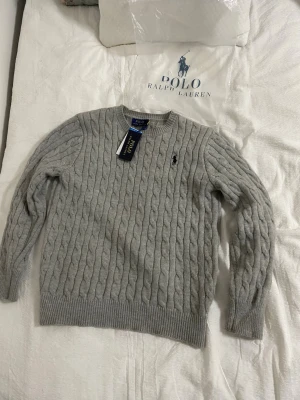Grå kabelstickad tröja Polo Ralph Lauren - Snygg grå kabelstickad tröja från Polo Ralph Lauren med rund halsringning och klassisk broderad logga på bröstet. Tröjan har ribbade muddar vid ärmslut och nederkant och är tillverkad i mjukt material som passar perfekt till höst och vinter. Packas ordentligt och skickas inom 24 timmar