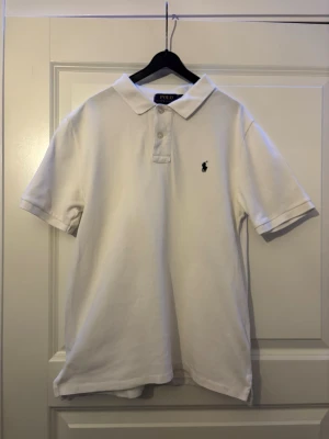 Ralph Lauren vit piké storlek 164 -  *   Färg: Vit *   Stil: Klassisk pikétröja *   Märke:  Ralph Lauren Polo     *   En traditionell pikékrage     *   Knäppning med två knappar     *   Broderad Polo-logotyp på bröstet *   Material: Bomull *   Passform: Designad för en normal passform *   Storlek: 164