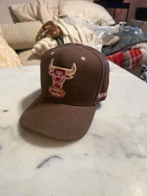 Brun Chicago Bulls keps Mitchell & Ness - Säljer en brun keps från Mitchell & Ness med Chicago Bulls-logga i rosa och vitt framtill. Kepsen har böjd skärm, broderade ventilationshål och NBA-märke baktill. Snygg streetwear-vibe och passar perfekt till en avslappnad stil.
