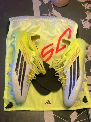 Adidas F50 elite fotbollsskor - Säljer ett par Adidas F50 fotbollsskor i en fräsch gul färg med svarta och röda detaljer. Skorna har klassiska tre ränder på sidan, snörning och en lätt, syntetisk ovandel. Sulan har fasta dobbar för spel på gräs. Medföljer gör en matchande skopåse och extra innersulor.