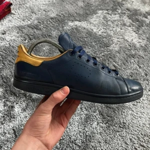 Adidas Raf Simons Stan Smith blå/gul - Snygga sneakers från Adidas och Raf Simons i mörkblått läder med gul hälkappa. Klassisk Stan Smith-silhuett med perforerat R på sidan, blå snörning och svart sula. Perfekta för dig som gillar stilrena men unika detaljer.✨ Det står storlek 44 men passar även de som har 45.           Vid eventuella frågor är det bara att skriva ✅ dem är lite slitna men har mycket kvar att ge. (Nypris runt 3500)
