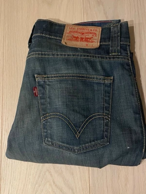 Blå Levi's jeans med raka ben - Säljer ett par klassiska blå Levi's jeans med raka ben och femficksmodell. Jeansen har tydliga kontrastsömmar, läderpatch bak i midjan och den ikoniska röda Levi's-taggen på bakfickan. Tillverkade i slitstark denim, perfekta för en avslappnad stil.