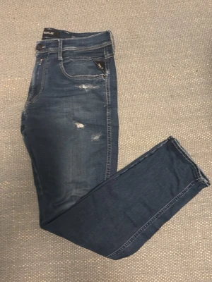 Mörkblåa jeans med slitningar från Replay - Anbass - Ett par blå jeans från Replay i modellen anbass med snygga slitningar i storlek 33/32. Jeansen är endast använda ett fåtal gånger. Säljer då jag växt ur jeansen, skriv om ni har några funderingar! Pris kan diskuteras.