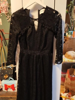 Svart spetsklänning från Pure Female - Svart långklänning från Pure Female med v-ringad hals och dekorativ spets över hela klänningen. Klänningen har trekvartsärm och ett litet nyckelhålsutskärning framtill. Perfekt för dig som gillar elegant och stilren design.