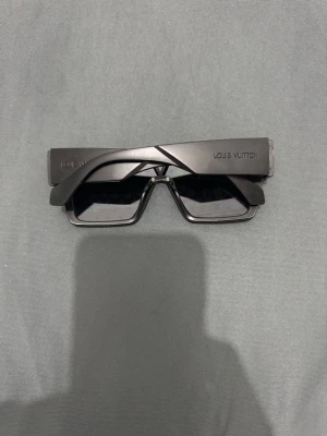 Svarta Louis Vuitton solglasögon - Säljer ett par svarta solglasögon från Louis Vuitton med futuristisk, rektangulär form och spegelglas med LV-monogram. Bågarna är breda och glasen har en cool, reflekterande finish. Perfekta för dig som vill sticka ut med en exklusiv accessoar.