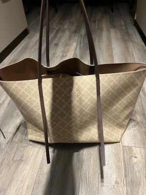 Malene birger väska i beige - Stilren shoppingväska i beige med diskret rutmönster och långa bruna axelband. Väskan är rymlig med öppen topp och har en praktisk innerficka. Det är den större versionen. Änvänd fåtal gånger