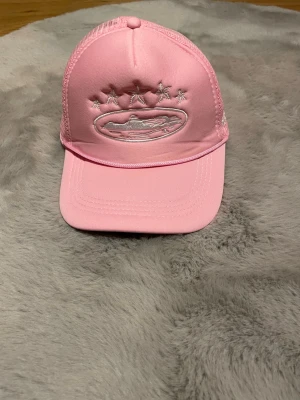 Rosa truckerkeps med broderi - Snygg rosa truckerkeps med vitt broderat motiv framtill och mesh på sidorna för extra ventilation. Kepsen har böjd skärm och justerbar snapback baktill. Perfekt för dig som vill sticka ut med en färgglad accessoar.