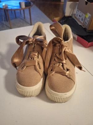 Beige sneakers från Go Sexy 37 - Stilrena beige sneakers från Go Sexy med bred platåsula och stora satinband som skosnören. Ovandelen är i mockaliknande material och insidan är fodrad med mjukt tyg. Bak på hälen finns vit detalj med guldfärgad logga.
