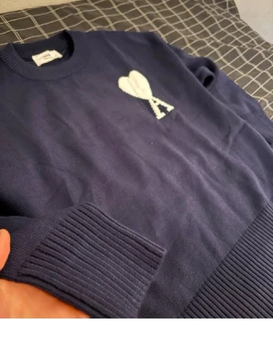 Mörkblå stickad tröja Ami Paris - Snygg mörkblå stickad tröja från Ami Paris med vitt hjärta och bokstaven A broderat på bröstet. Tröjan har rund halsringning, ribbade muddar och nederkant. Perfekt för dig som gillar stilrena detaljer och klassisk design.