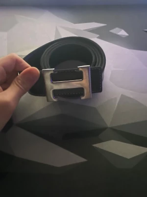 Svart bälte med silverfärgat H-spänne - Snyggt svart bälte med ett stort, silverfärgat H-format spänne. Bältet har en stilren och modern look som passar till många outfits. Perfekt accessoar för att lyfta din stil.