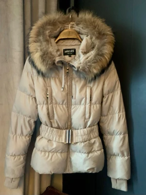 Beige dunjacka med pälskrage Hollies - Snygg beige dunjacka från Hollies med stor fluffig pälskantad huva och bredt bälte i midjan med metallspänne. Jackan har dragkedja, fickor med dragkedja och ribbade muddar vid ärmslut. Perfekt för kalla vinterdagar.