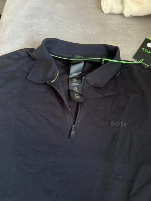 Svart polotopp från BOSS med zip - Snygg svart polotopp från BOSS med klassisk krage och halv dragkedja framtill. Diskret BOSS-logga på bröstet och en liten grön detalj vid kragen. Tillverkad i ett mjukt och bekvämt material som passar perfekt till många olika stilar.